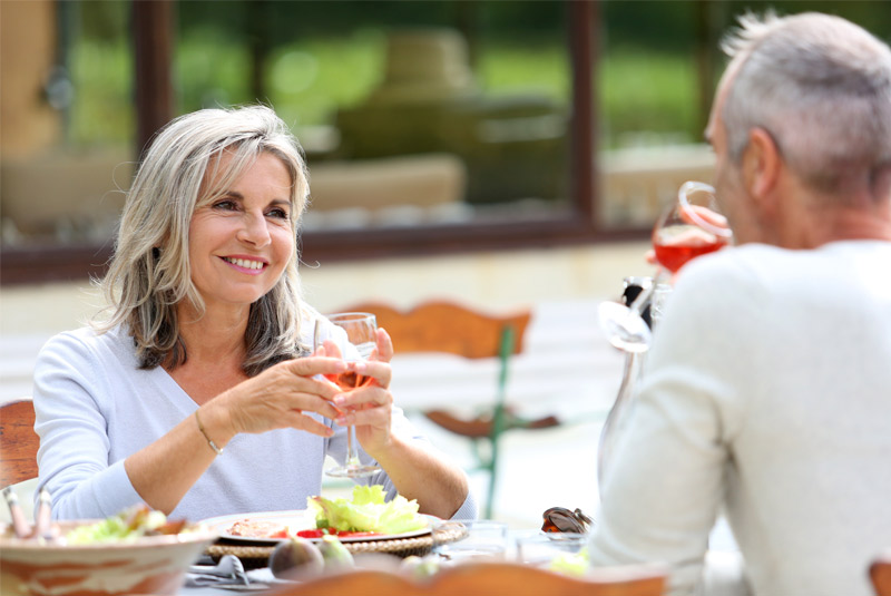 Lady with white hair drinking wine at a table eating dinner also because she went to Restore Denture and Implant Center for her loose denture